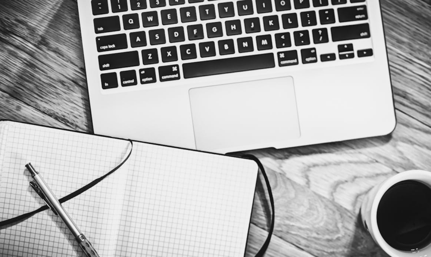 A laptop near a notebook, pen, and a cup of coffee